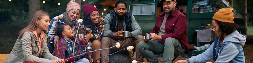 Multiracial families roasting marshmallows while gathering around campfire in woods.